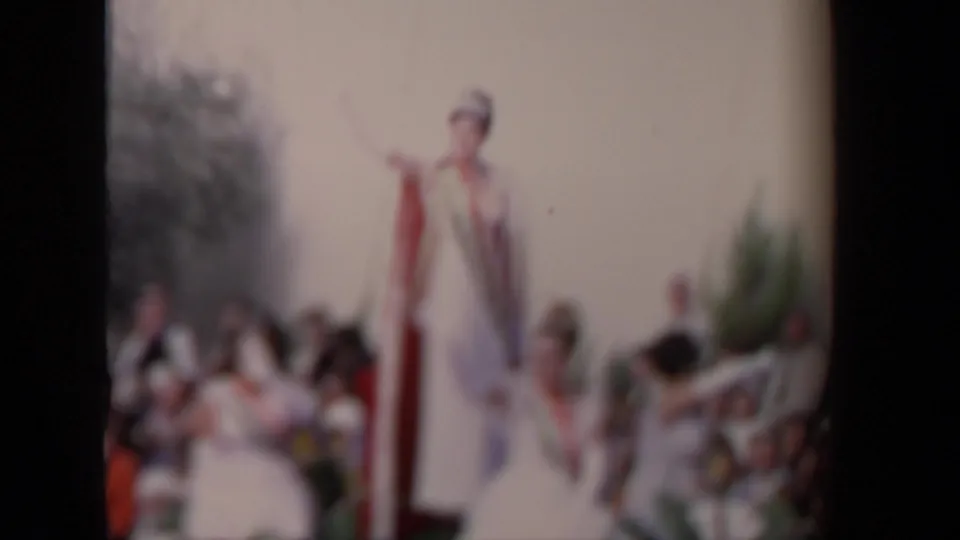 Soft-focus 1967 Los Angeles parade frame with a standing performer in white attire on the float and spectators behind.