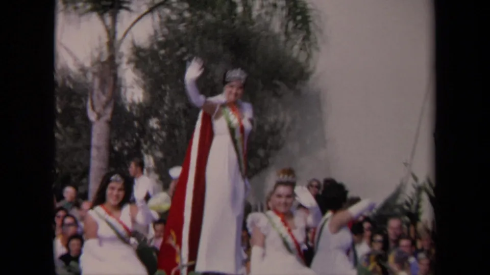 Home-movie frame of a 1967 Los Angeles parade queen waving in white dress and tiara with attendants and spectators around her.