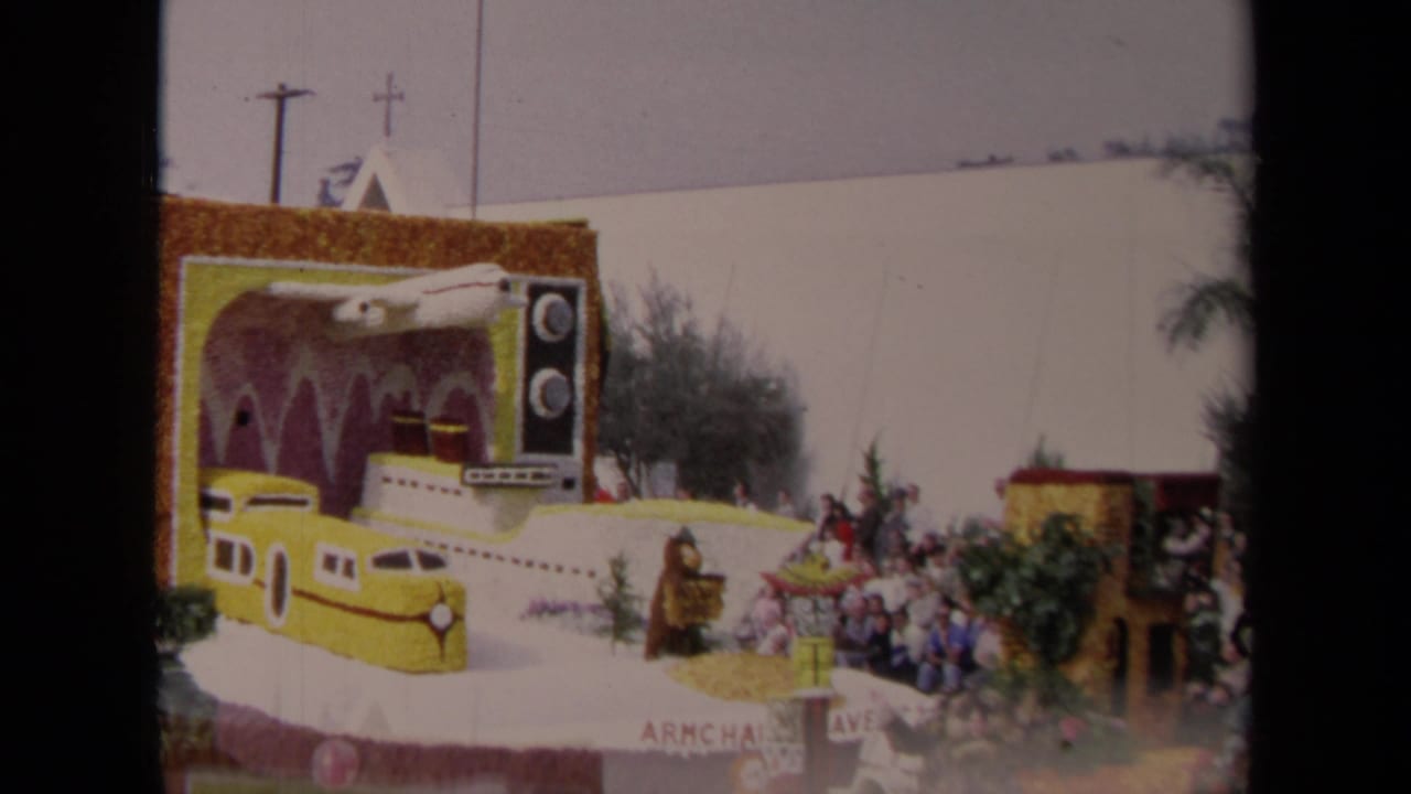 1967 Los Angeles 8mm frame showing a decorated parade float with a yellow train, submarine-like body, oversized television prop, and curbside spectators.