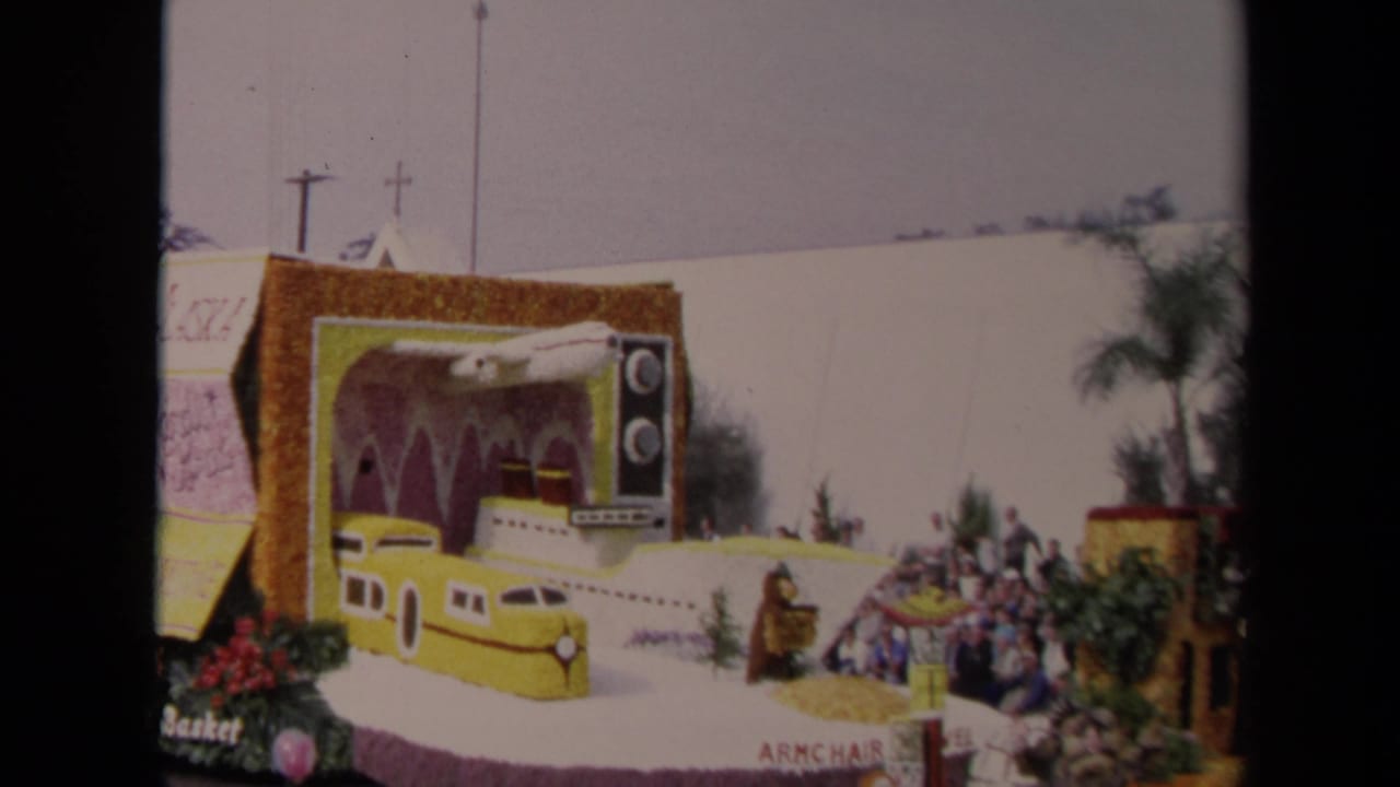 Parade float with yellow train and television prop passing a seated crowd near palm trees.