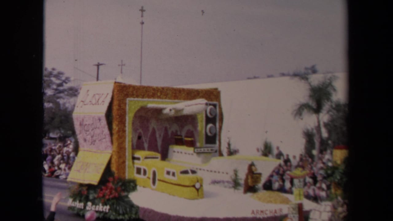 1967 Los Angeles home-movie frame showing the side of a parade float with a television prop, yellow transport design, flowers, and spectators.
