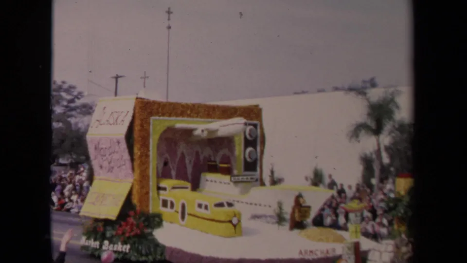 1967 Los Angeles home-movie frame showing the side of a parade float with a television prop, yellow transport design, flowers, and spectators.