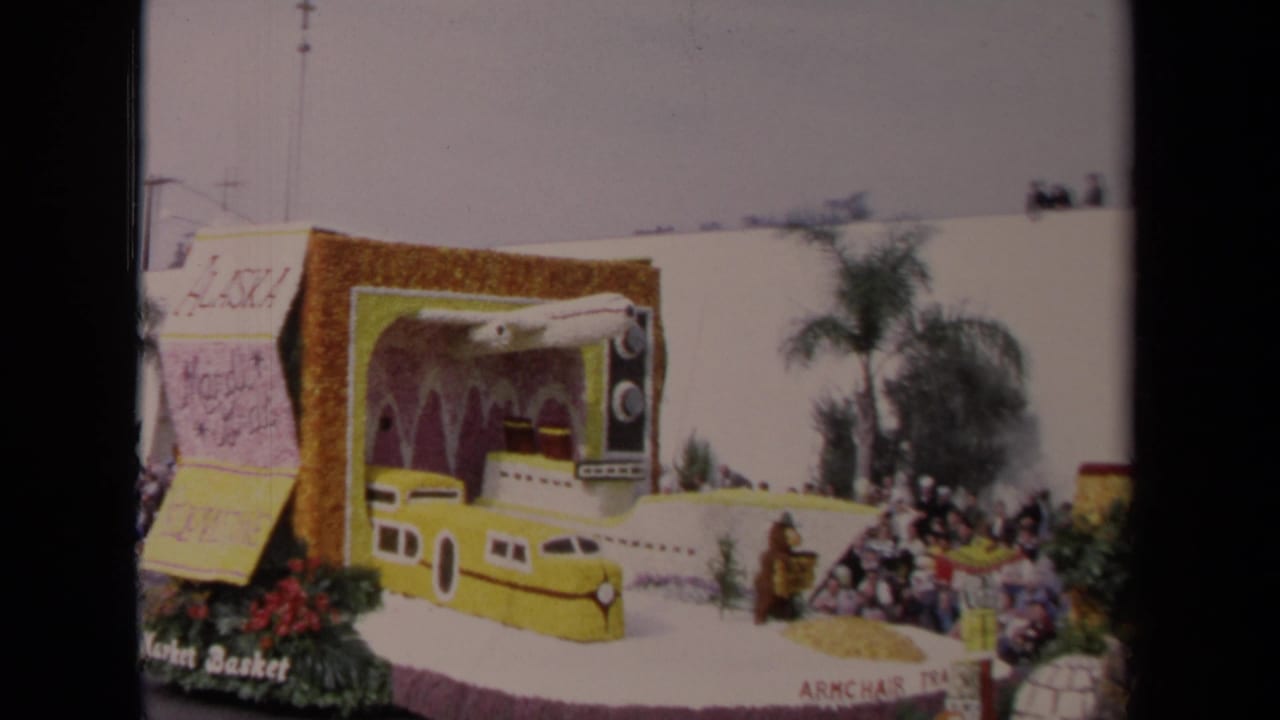 1967 Los Angeles 8mm frame of a decorated parade float with a yellow train-like front, large television prop, flowers, and curbside viewers.