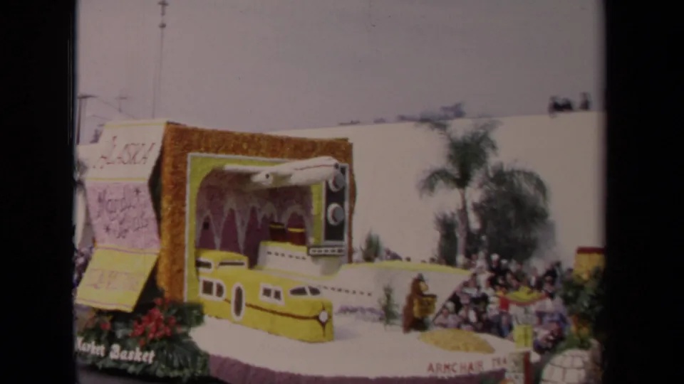 1967 Los Angeles 8mm frame of a decorated parade float with a yellow train-like front, large television prop, flowers, and curbside viewers.