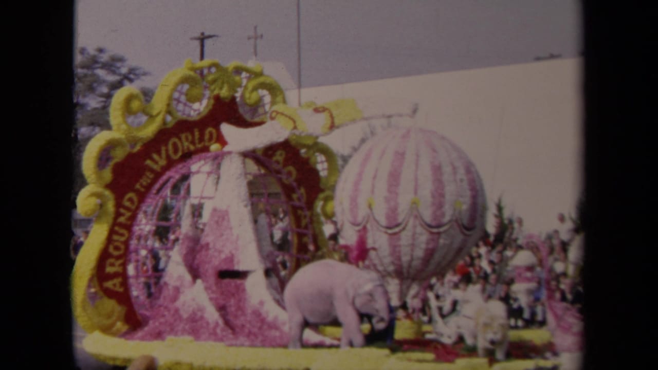 1967 Los Angeles parade float with Around the World lettering, a striped balloon prop, pink elephant figures, floral trim, and a crowd behind it.