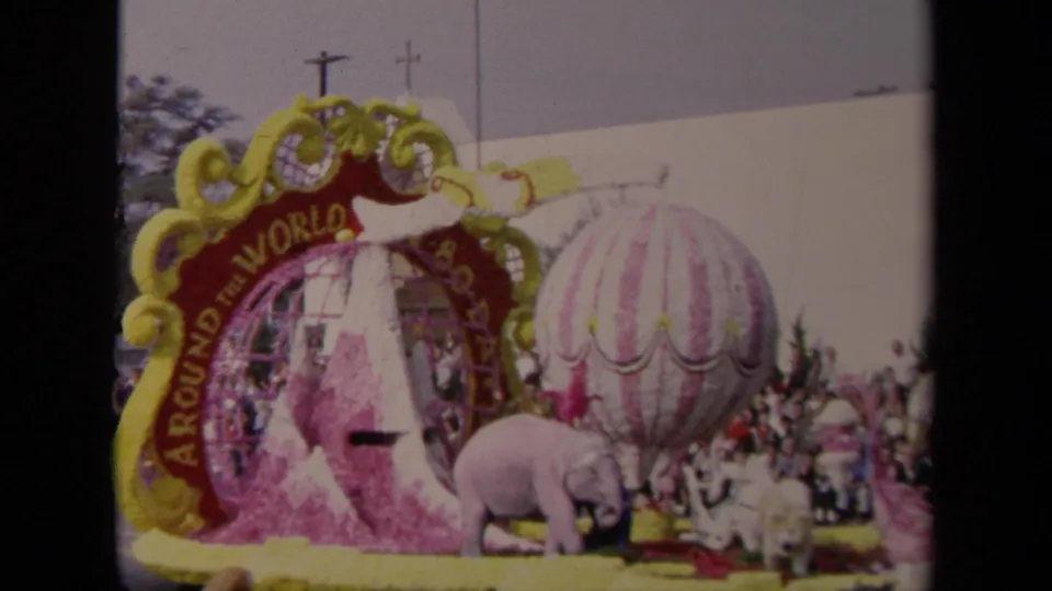 1967 Los Angeles parade float with Around the World lettering, a striped balloon prop, pink elephant figures, floral trim, and a crowd behind it.