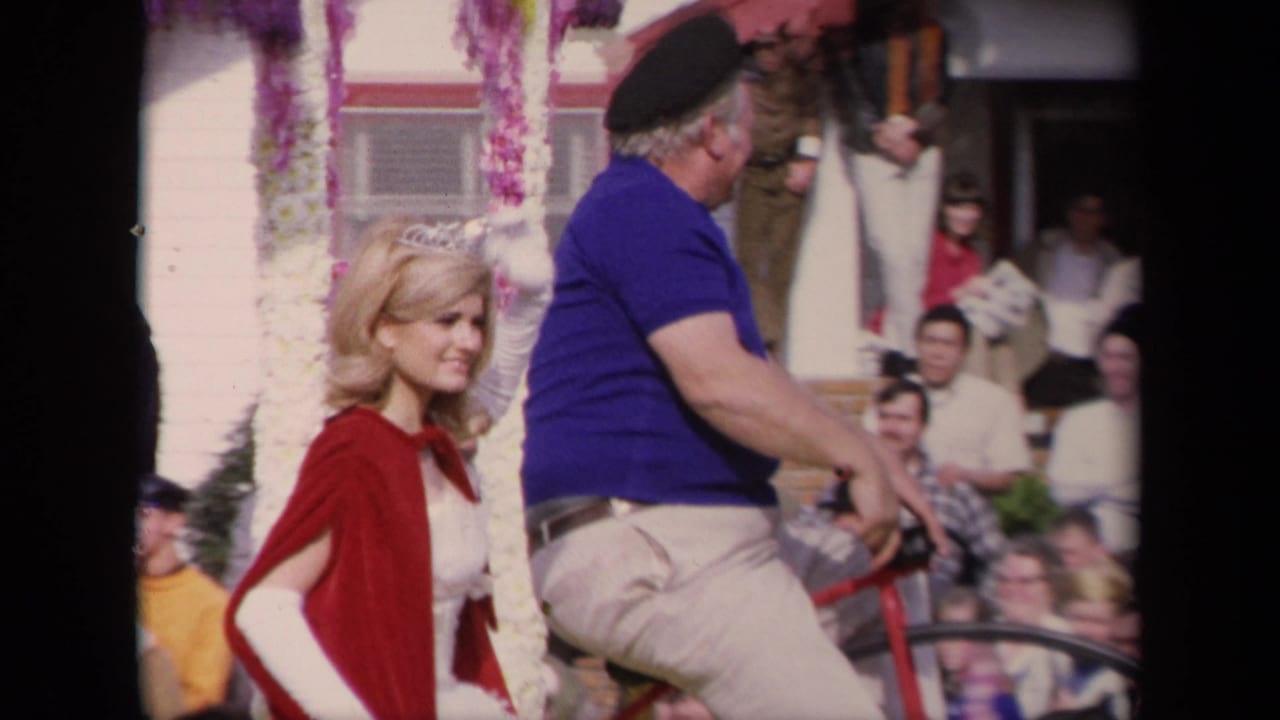 Woman in red cape and tiara beside a man in blue shirt and beret on a bicycle during a parade.