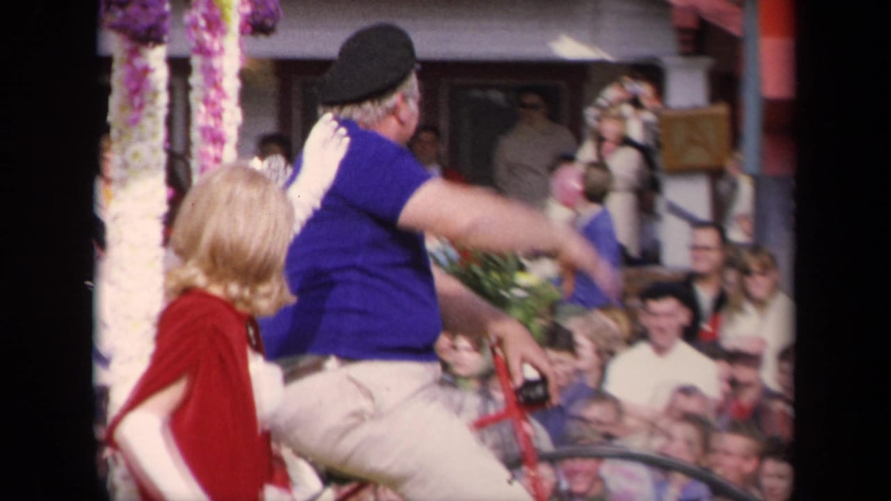 Side view of the 1967 Los Angeles parade with a man in a blue shirt on a bicycle beside a woman in a red cape, with spectators in the background.