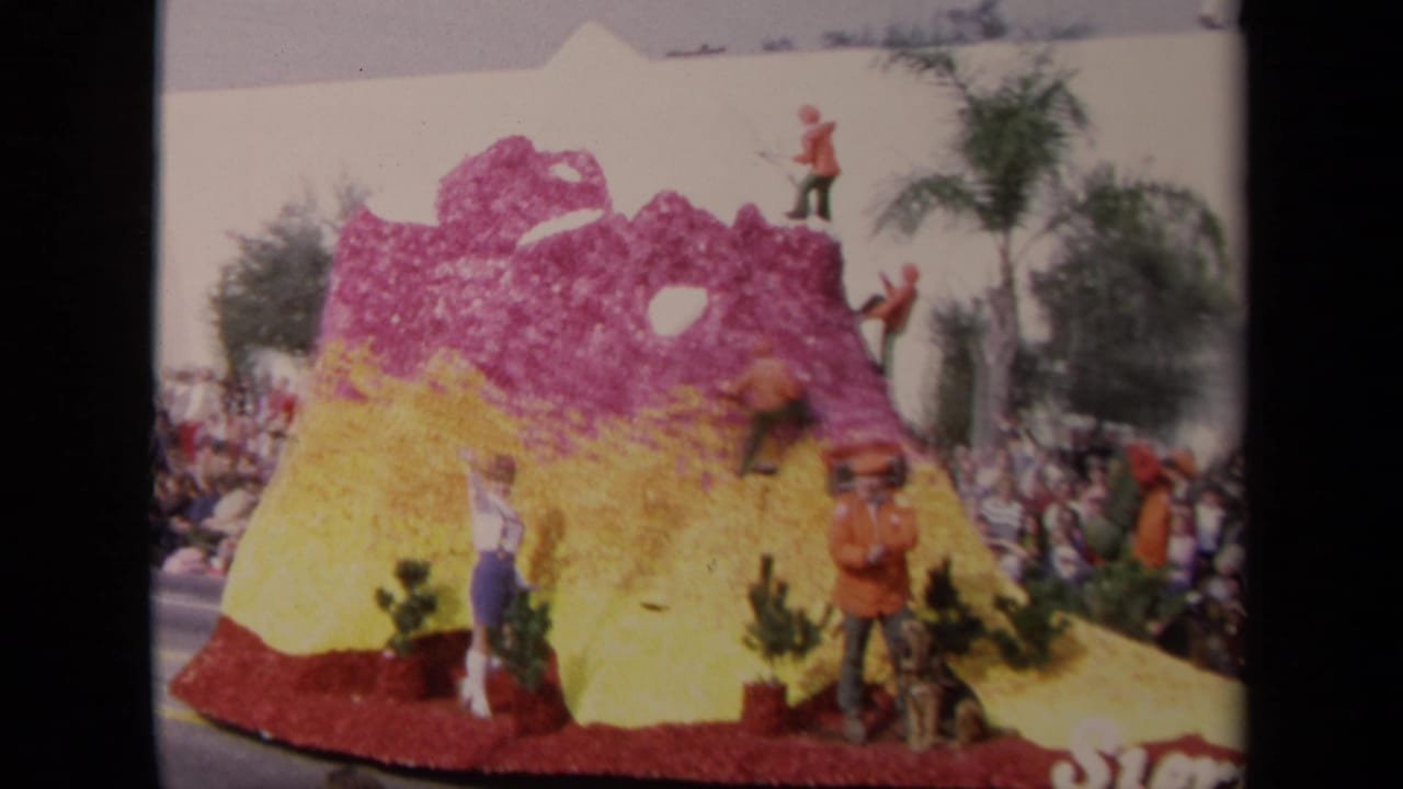 Colorful floral parade float shaped like a mountain with spectators along the street.