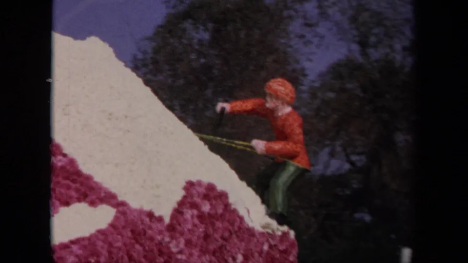 The 8mm frame shows a climber figure posed on a pink and white floral float surface with trees in the background.