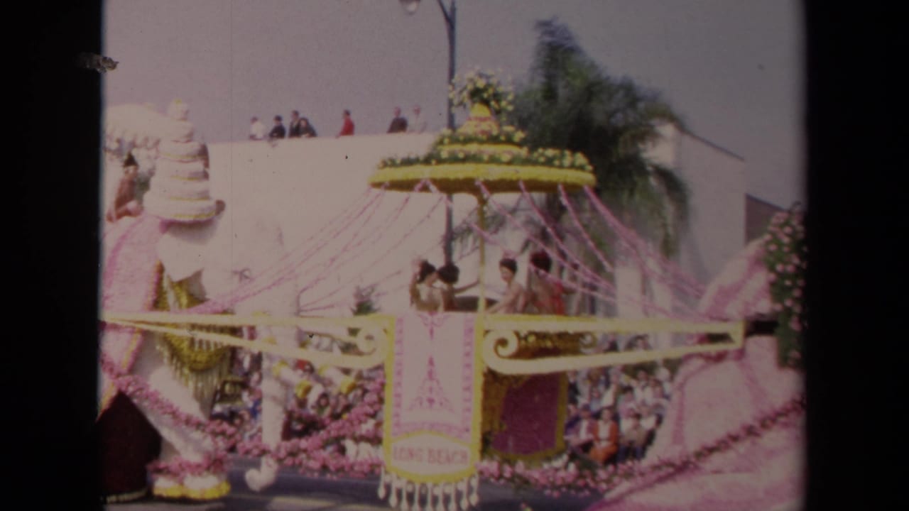1967 Los Angeles home-movie frame showing a side view of the decorated elephant float with riders, pink streamers, and spectators behind it.