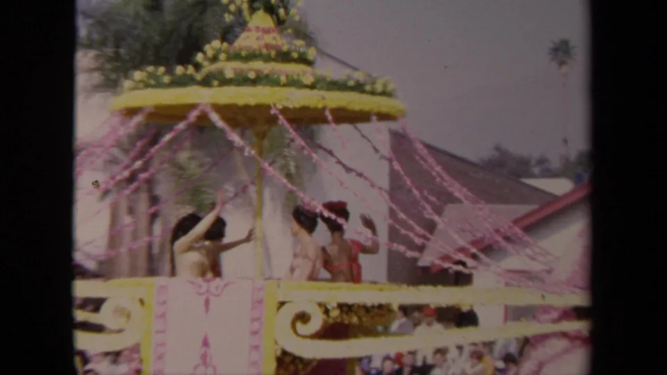 1967 Los Angeles parade frame focused on costumed riders beneath a yellow floral canopy, with pink streamers and spectators visible below.