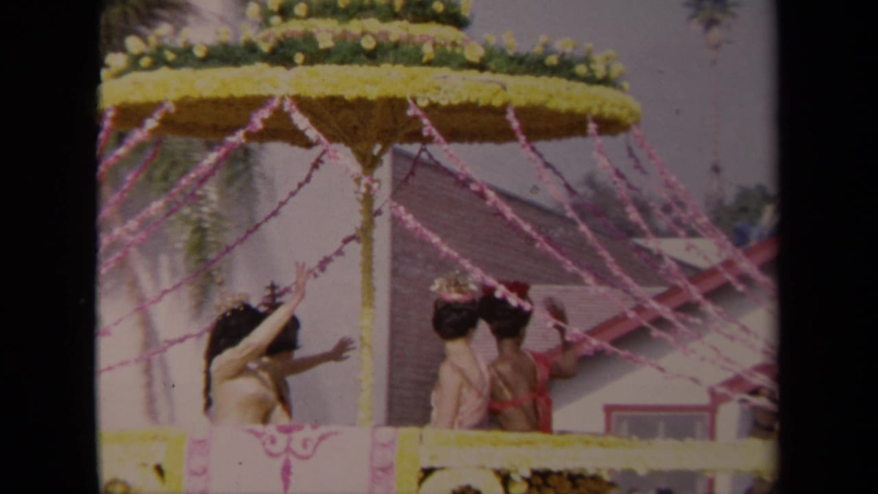 1967 Los Angeles home-movie frame showing women on a decorated parade float under a floral umbrella and pink streamers.