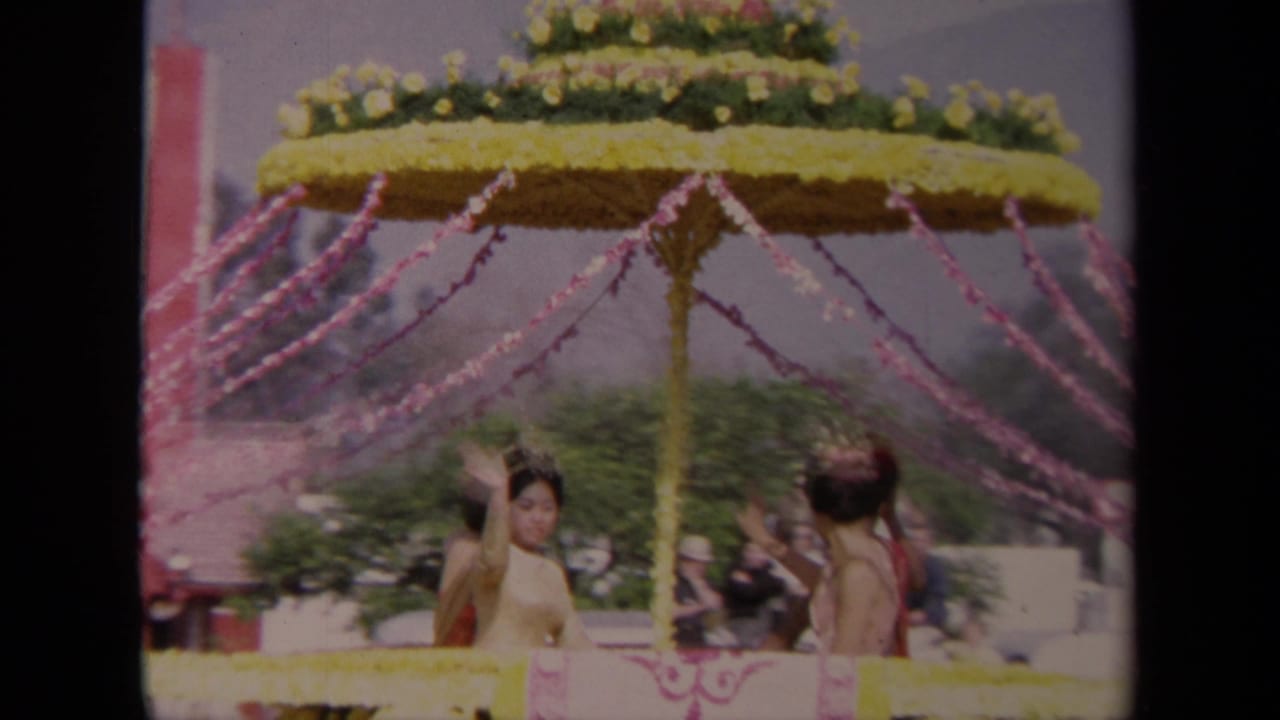 Costumed women ride on a float with a large flower-covered umbrella.