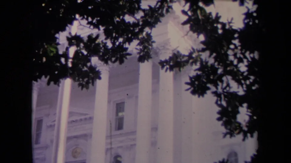 1967 home-movie frame showing a white columned exterior with a partial entrance area behind dark tree cover.