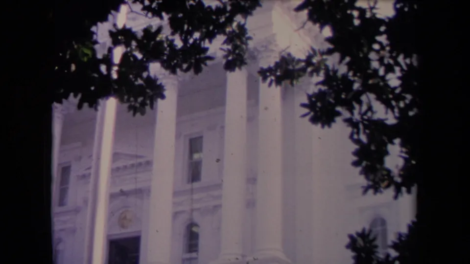 Muted 8mm frame of a columned white building exterior, with foliage obscuring part of the facade and doorway.