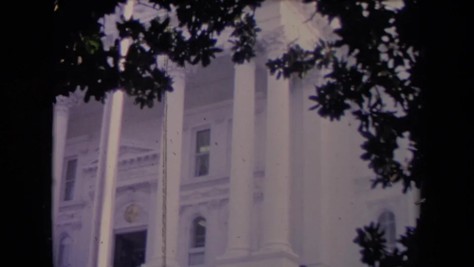 1967 8mm frame of a grand white neoclassical facade with columns, a flagpole, windows, and tree silhouettes in front.