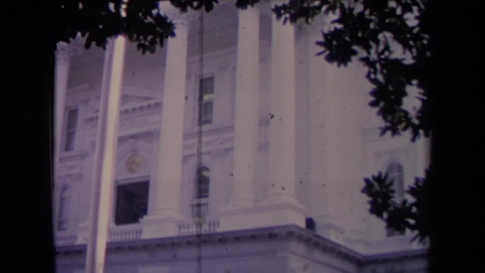 1967 8mm frame of a white civic-style building facade, with tall columns, flagpole, lower arches, and dark foliage framing the view.