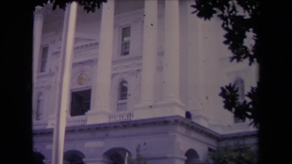 1967 8mm frame showing a tree-framed white public building facade with tall columns, arched lower openings, and visible film grain.