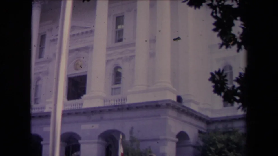 8mm frame of a formal white building with columns and arches, seen through trees above people gathered outside.