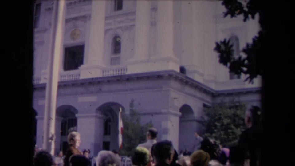 Lower-framed 8mm view showing more of the crowd below the columned building and a flag near the entrance.