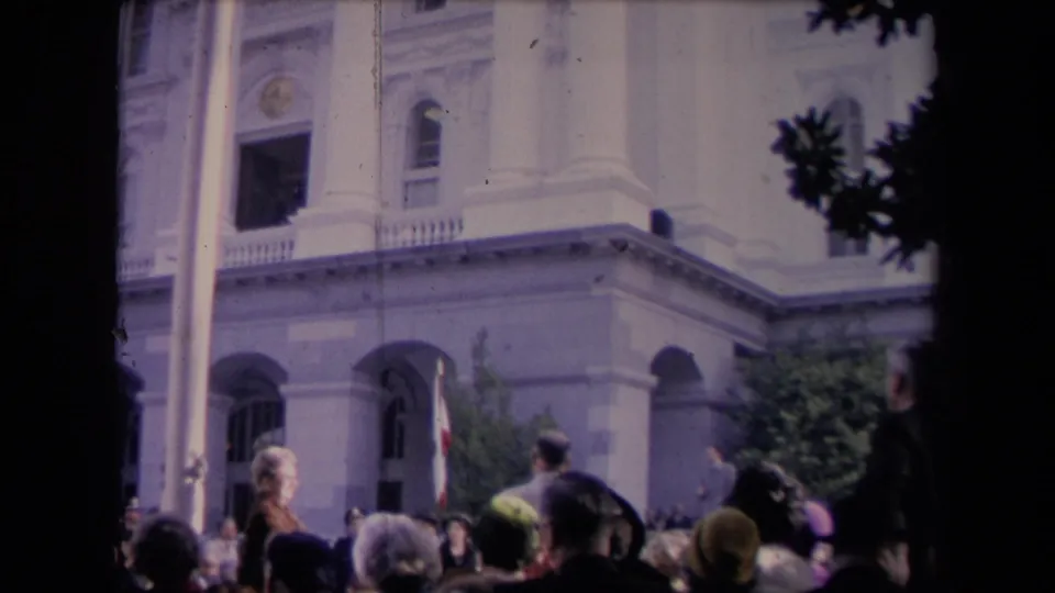 Final frame of spectators gathered near a white columned building, with branches and a dark trunk framing the scene.