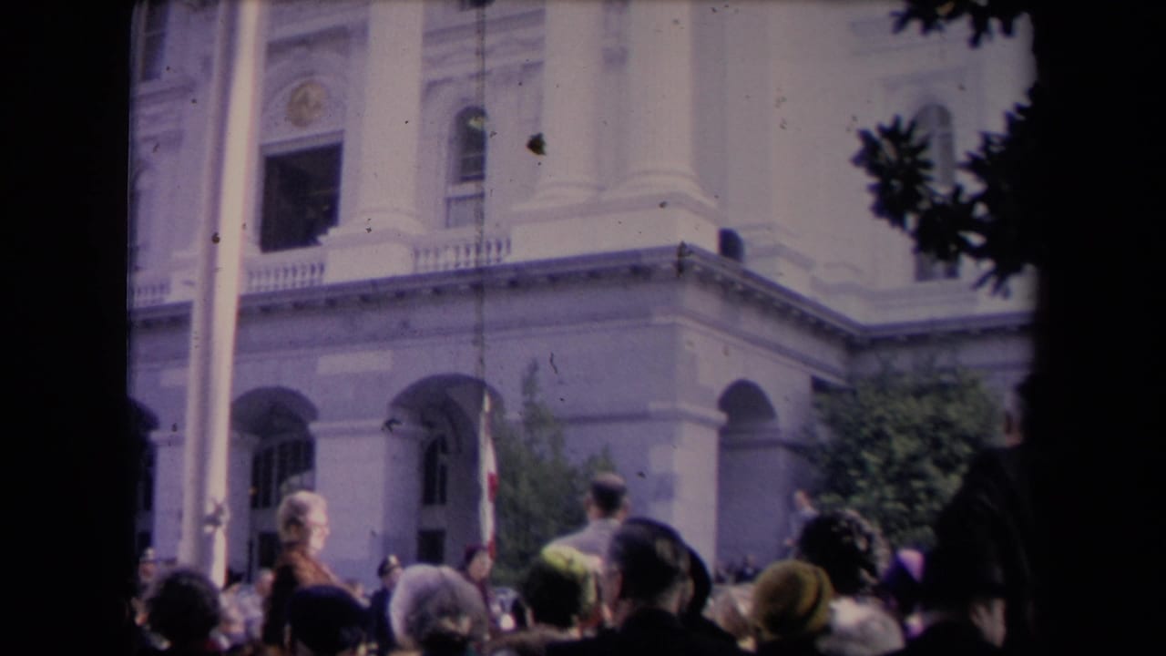 Crowd gathered before a columned public building with a flagpole and trees.