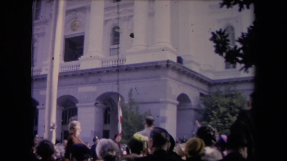 Spectators stand outside a classical building during a public political gathering, filmed from within the crowd on 8mm.