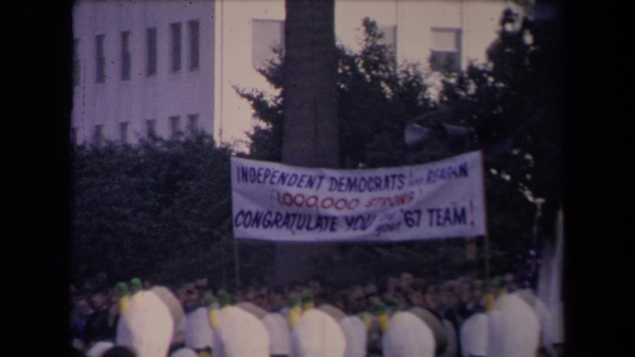 Large political banner above a crowd near trees and a building facade.