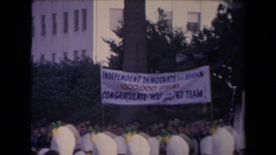 A banner for Independent Democrats is held above a crowd, with trees and a public building visible behind it.
