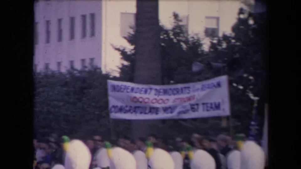 The camera frames a political banner over a dense crowd, with soft focus and faded 8mm color.