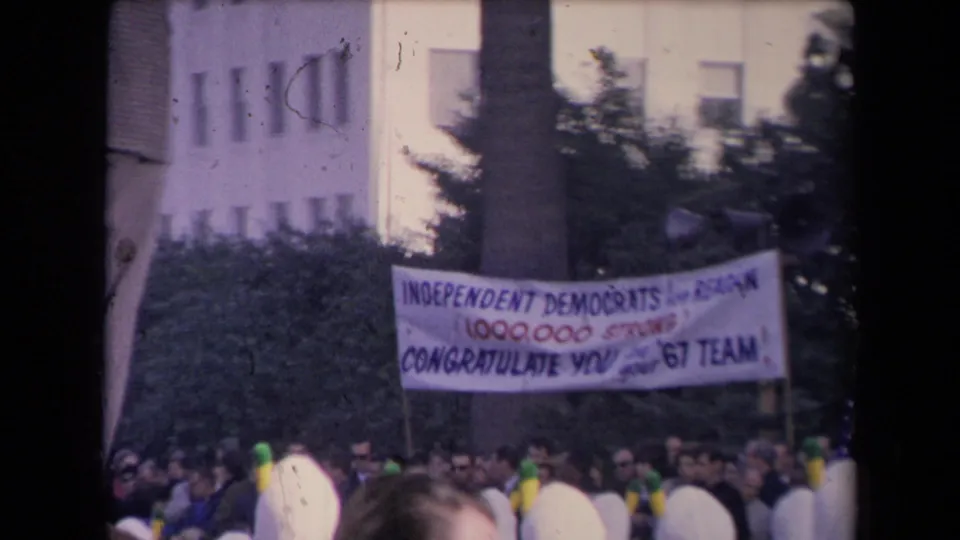 A crowd watches beneath an Independent Democrats banner, with a building and trees behind the gathering.