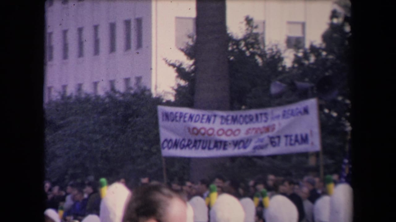 Crowd beneath a political congratulatory banner with a building and trees behind it.