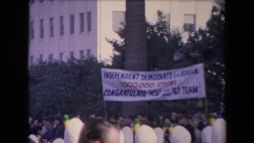 Outdoor crowd in archival 8mm footage beneath a banner congratulating a political team, with trees and a civic-looking building in the background.