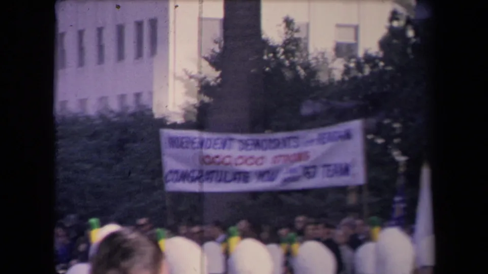 Archival home-movie frame showing a political banner above a packed outdoor crowd, with foliage and a building visible behind it.