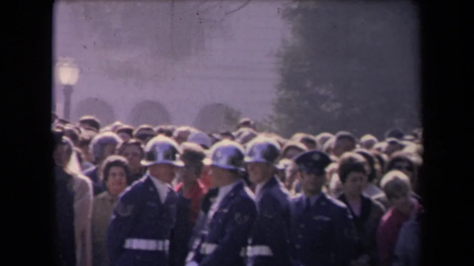 Uniformed guards with reflective helmets stand among spectators at a crowded outdoor political event, with haze and a lamp post behind them.