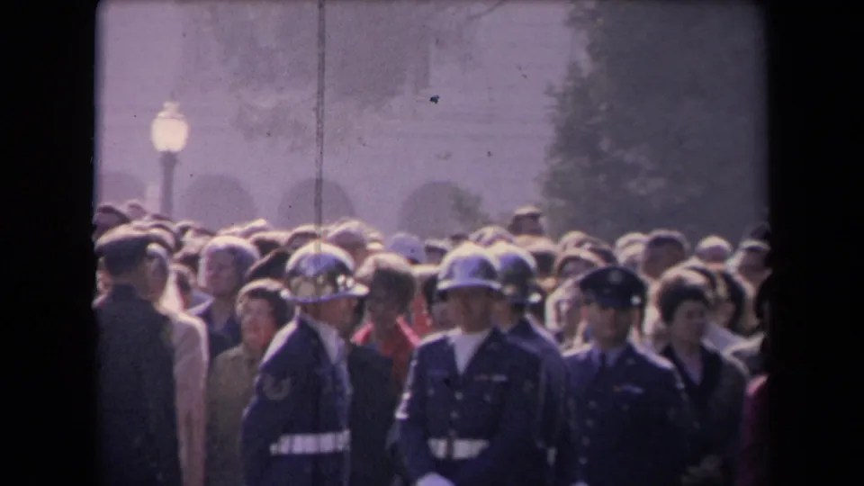 Archival 8mm frame of helmeted uniformed guards standing near spectators in a bright, hazy outdoor political crowd.