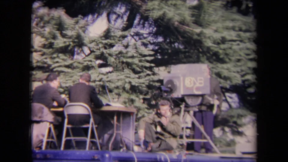 Outdoor 8mm view of men seated at a desk on a platform, with a large television camera and crew equipment beside evergreen trees.