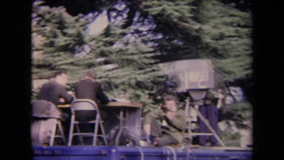 Home-movie frame showing a raised media platform with seated men, a bulky television camera, folding chairs, and evergreen trees in the background.
