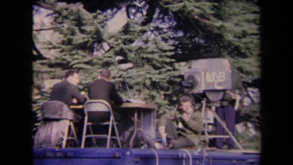 8mm frame of an outdoor media platform, with suited men at a desk and a large television camera marked with a visible number beside them.