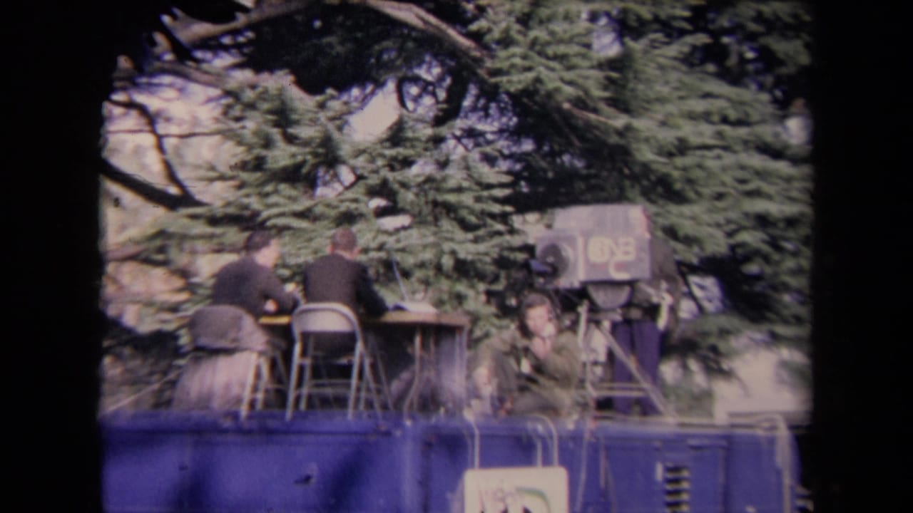 Broadcast crew seated on a raised mobile news platform beneath trees.