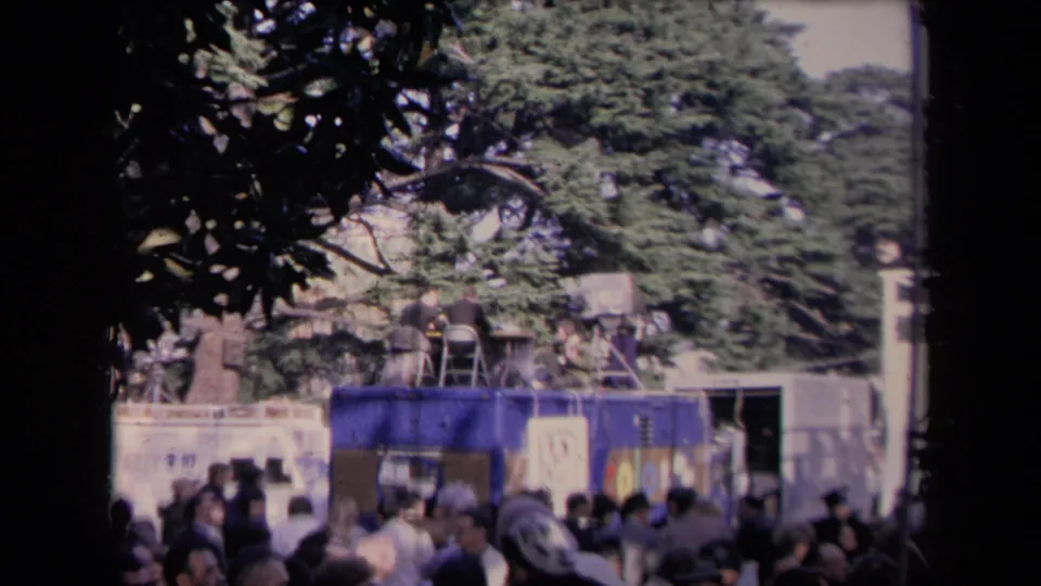 Handheld 8mm view of a crowd and raised news unit partly framed by tree branches at the Sacramento political event.