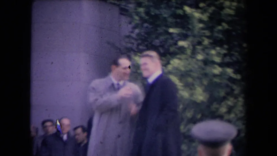 Soft-focus 8mm frame of suited men near a stone wall, with seated attendees behind them at the 1967 Sacramento political gathering.