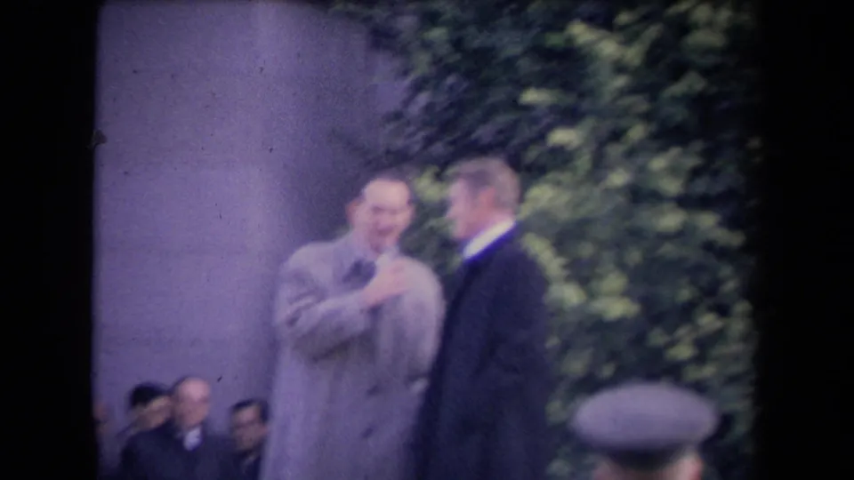 Soft 8mm frame of two formally dressed men conversing outdoors near a stone wall and foliage, with several onlookers seated behind them.