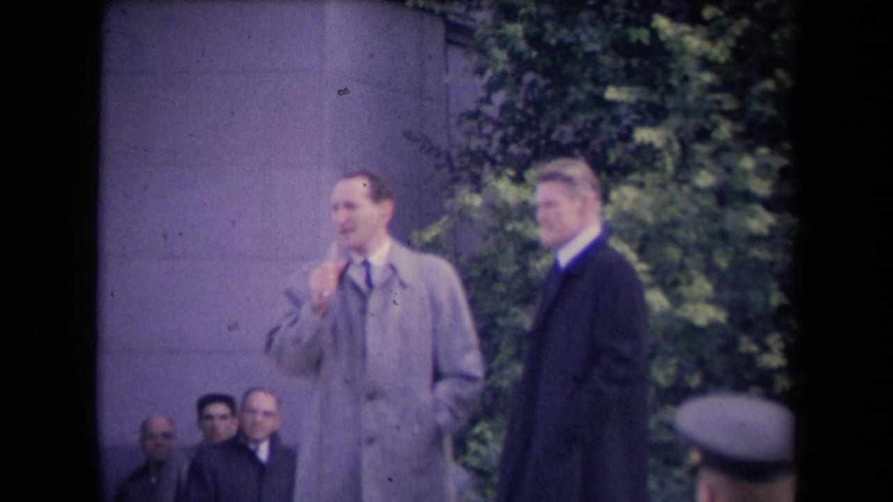 A man in a light overcoat and tie gestures near his face while a second man in a dark coat faces the camera, with seated observers behind them.
