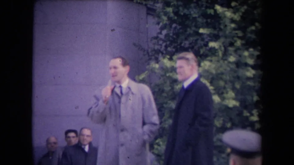 A man in a light overcoat and tie gestures near his face while a second man in a dark coat faces the camera, with seated observers behind them.