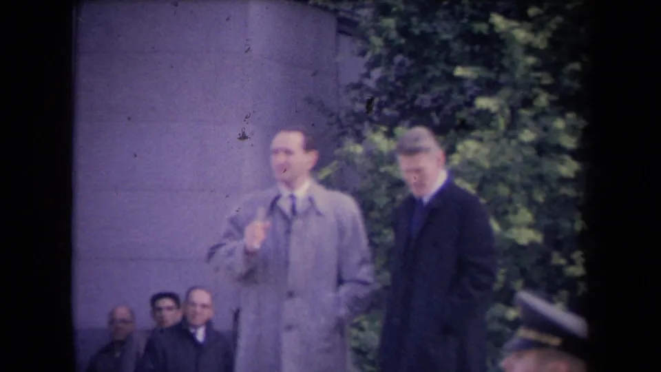 Outdoor 8mm frame showing a light-coated man gesturing as a dark-coated man looks downward, with seated men and foliage in the background.
