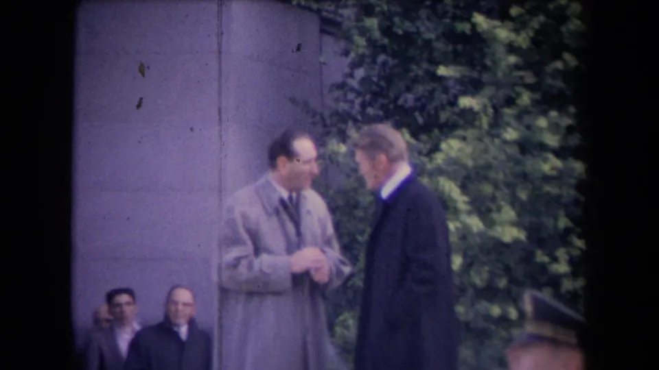 Two formally dressed men face each other closely in an outdoor public setting, with a stone wall, foliage, and seated observers behind them.
