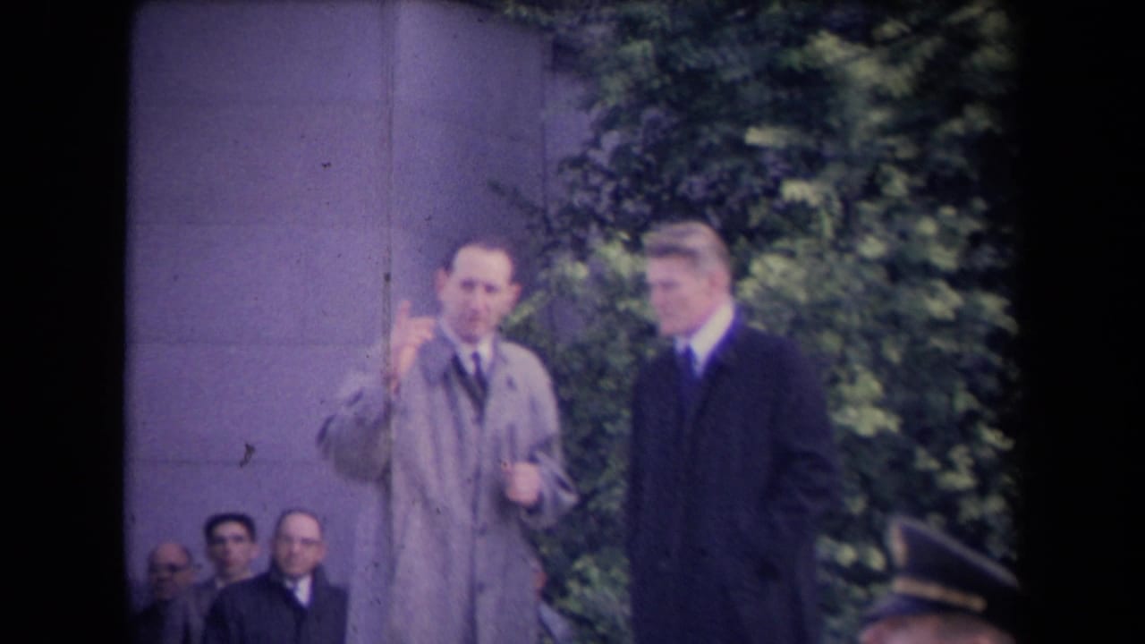 Two men in overcoats stand near a stone column with seated onlookers behind them.