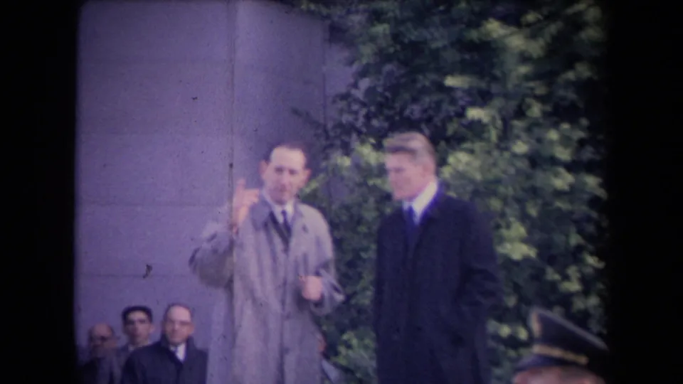 At a 1967 Sacramento political gathering, two formally dressed men stand before seated attendees beside a stone column and greenery.